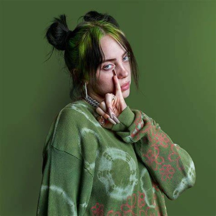 Billie Eilish Wearing a green top with jewelry in her ears and neck and round black spectacles.